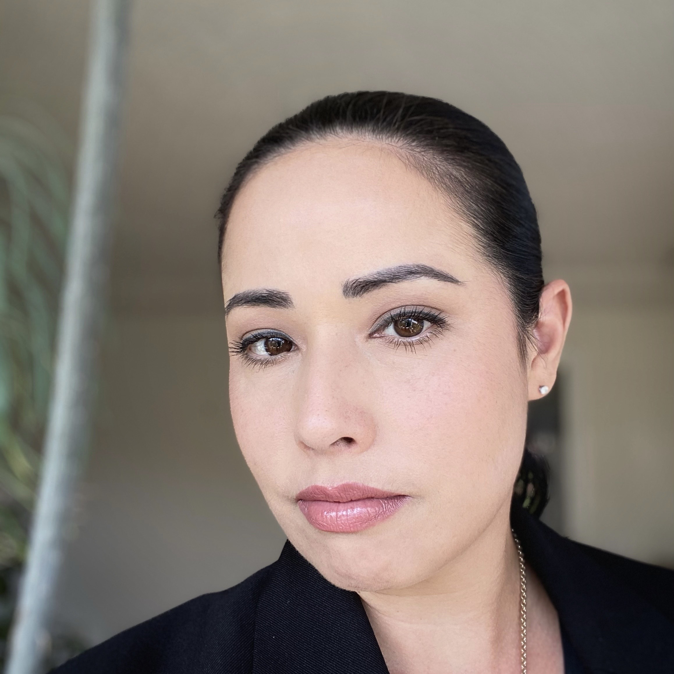A woman with fair skin looks serenely at the camera, her pink lips gently pressed together. Her black hair is pulled back. Dark eyebrows arch over her deep brown eyes. She wears a black jacket with a silver chain and earrings. A tree and beige surroundings are visible in the background.