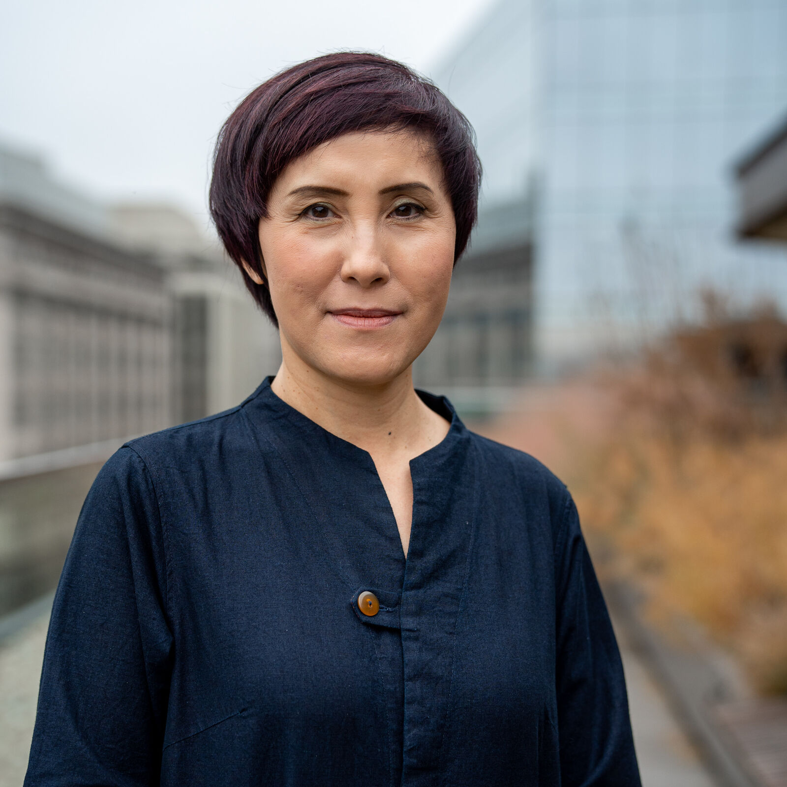 A Vietnamese woman stands regally in front of a cityscape flecked with the light browns of drying plants. She wears a navy blouse closed with a brown button with a small v at the neckline. A small smile plays on her lips.