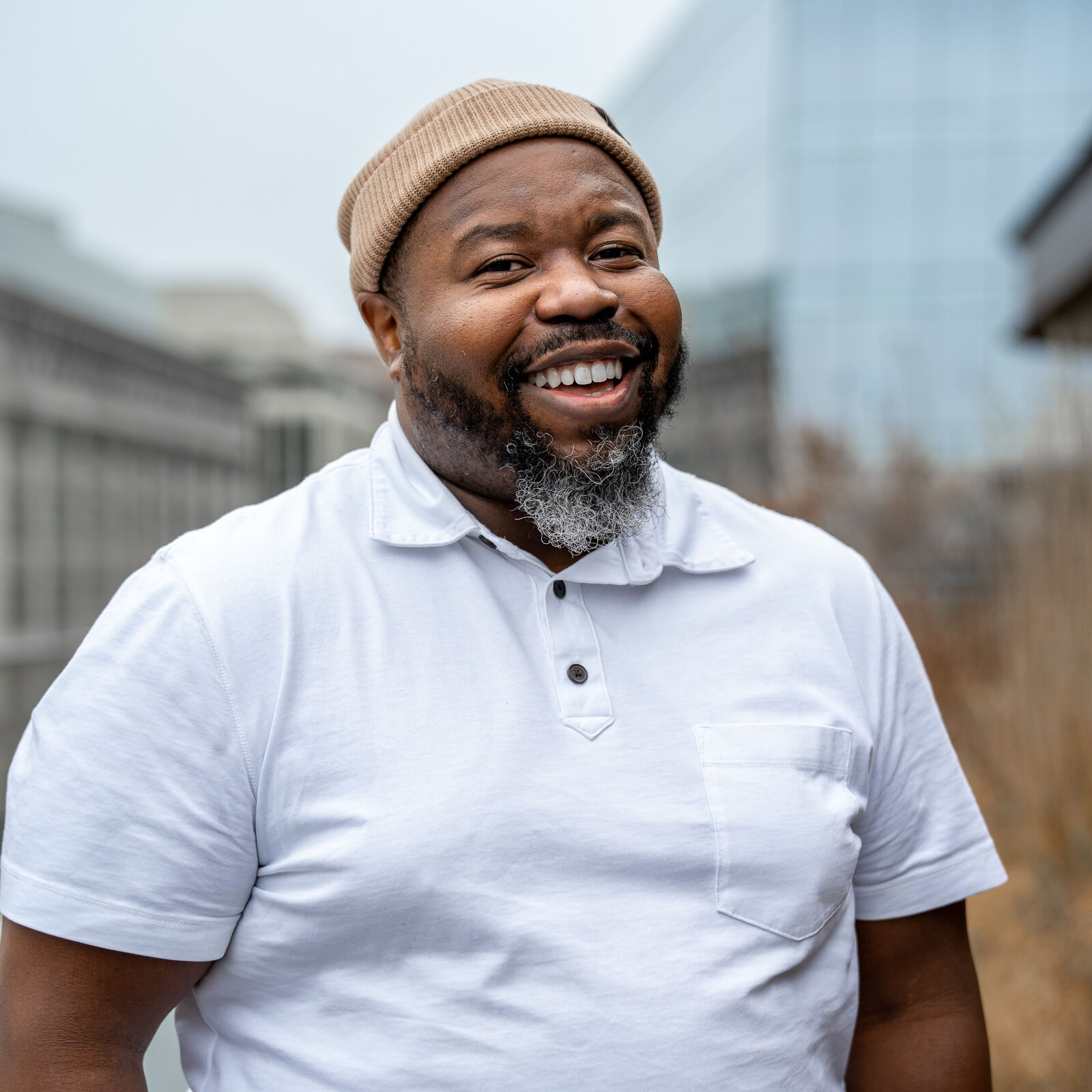 A Black man grins at the camera, his cheeks rounding with the smile as his white teeth drawout the colors of his white polo and the silvery flecks at the bottom of his black beard. He wears a tan beanie. A blurred cityscape lies behind him.