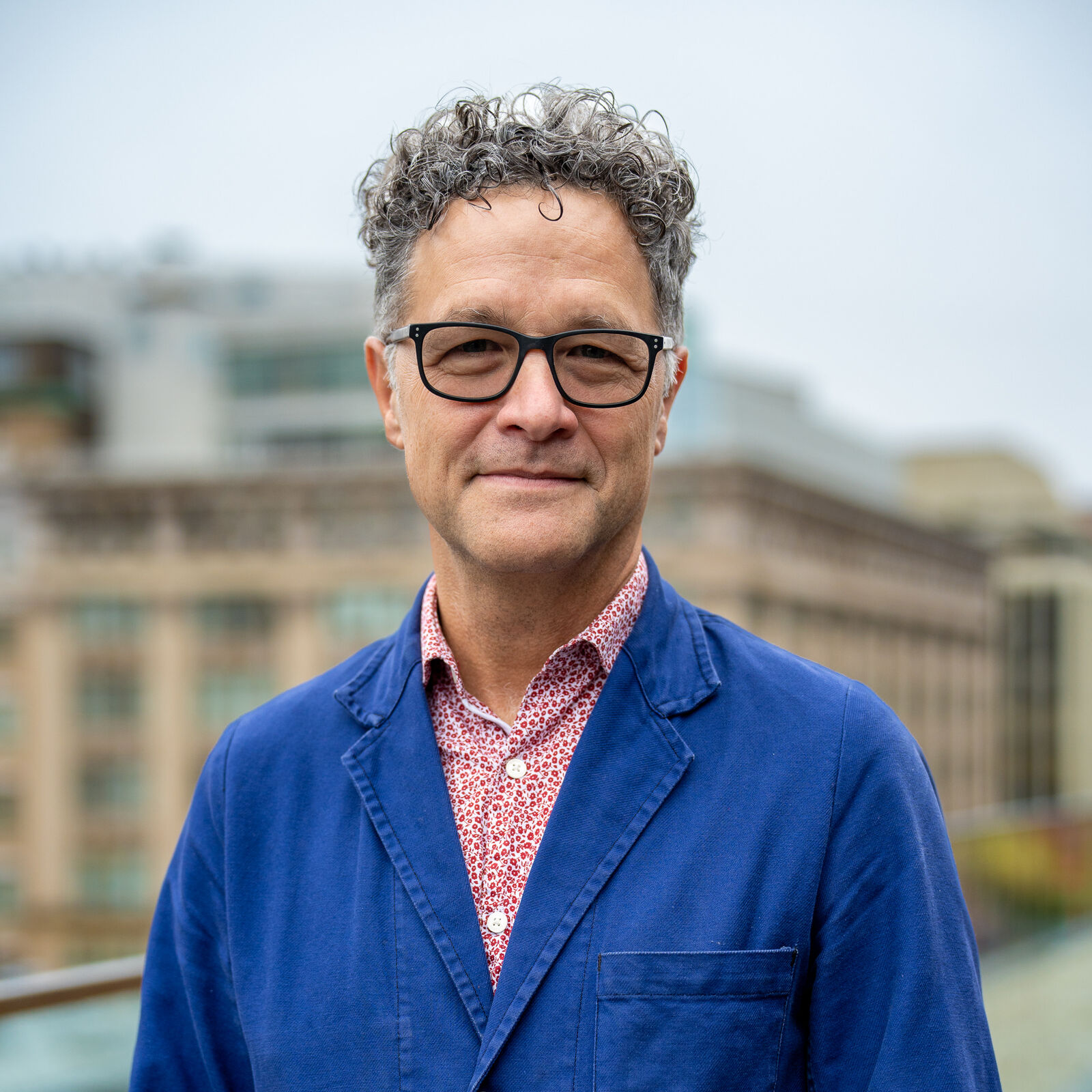 A man smiles at the camera, his lips pressed into a flat smile showing mirth at the corners. He wears large black-rimmed glasses with frames shaped like softened rectangles. His short hair curls atop his head in a mix of silver and dark gray. He wears a blue jacket with a red and white shirt with such a small pattern it appears to be pink. A cityscape in beige and gray is behind him.