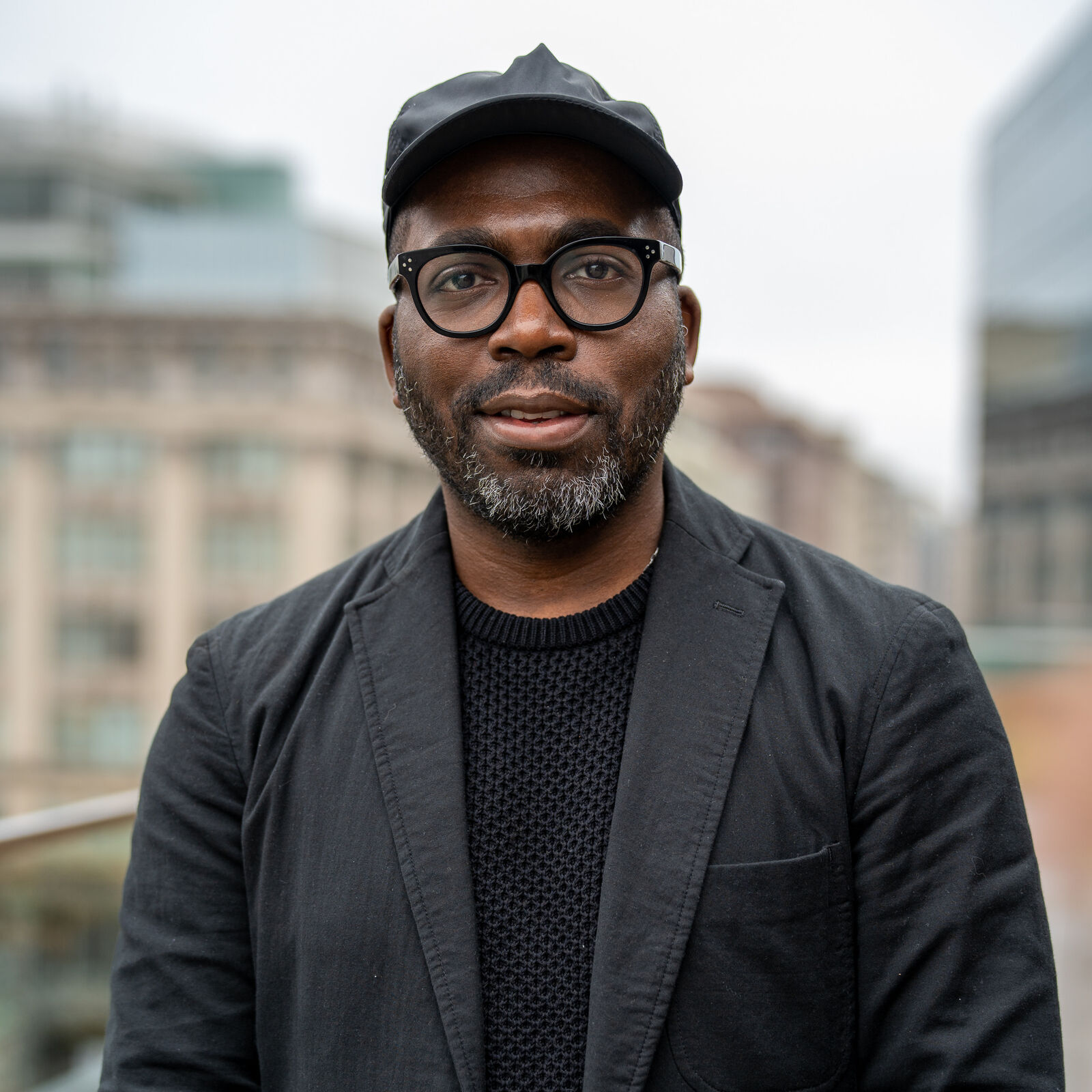 A Black man gazes into the camera, composed but still relaxed. He wears a black shirt with a slightly lighter black blazer and a black cap, as well as rounded black glasses. The clean lines of the frames bring out the colors in his beard: black with flecks of silver at the front. A cityscape is distantly visible in the background.