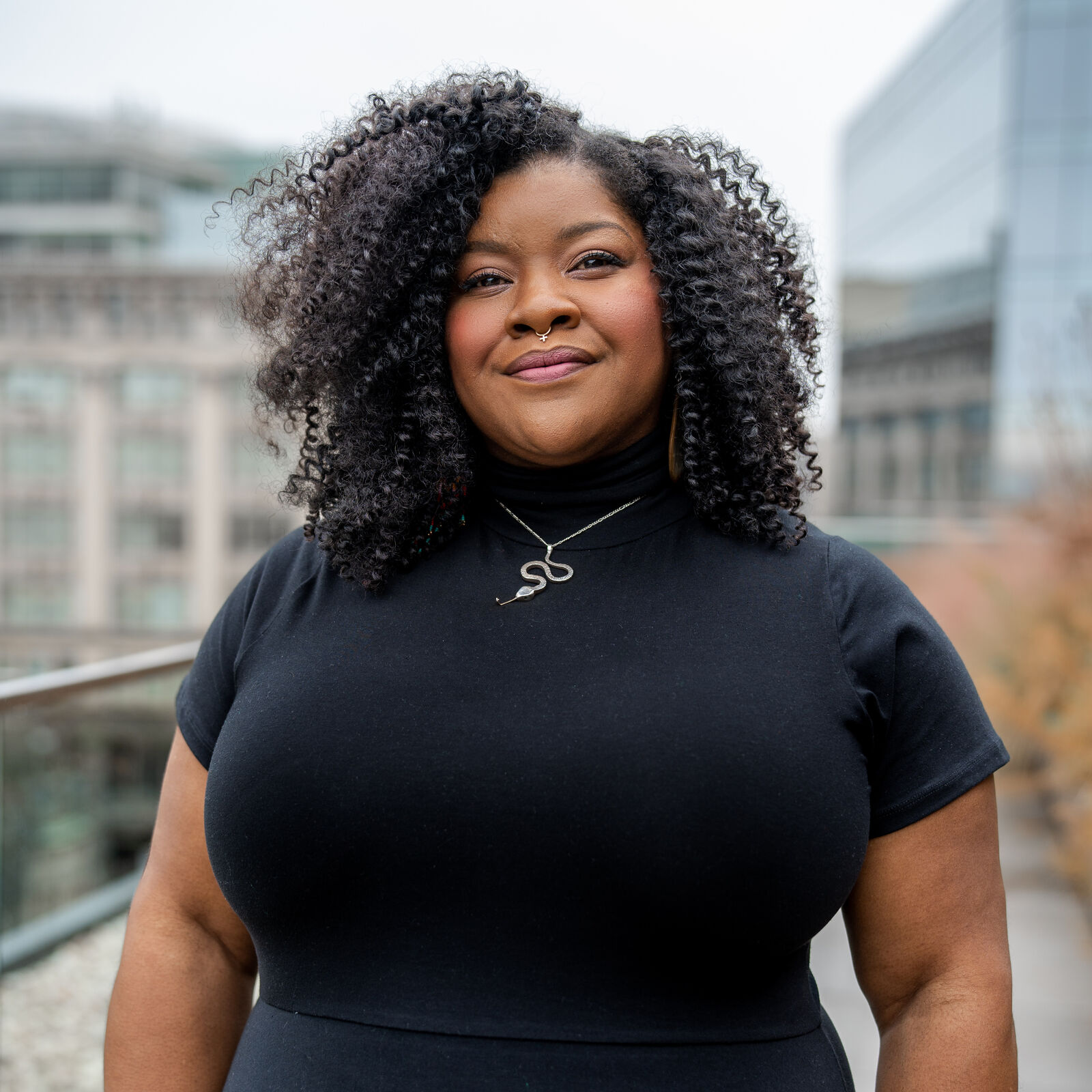 A Black woman smiles confidently, revealing rosy cheeks and pink lips. Her black hair curls in tight spirals around her head. A small silver piercing crosses the bottom center of her nose, and she wears a black dress with a silver serpent necklace. A cityscape with autumn tones and a gray sky is blurry but visible in the background.